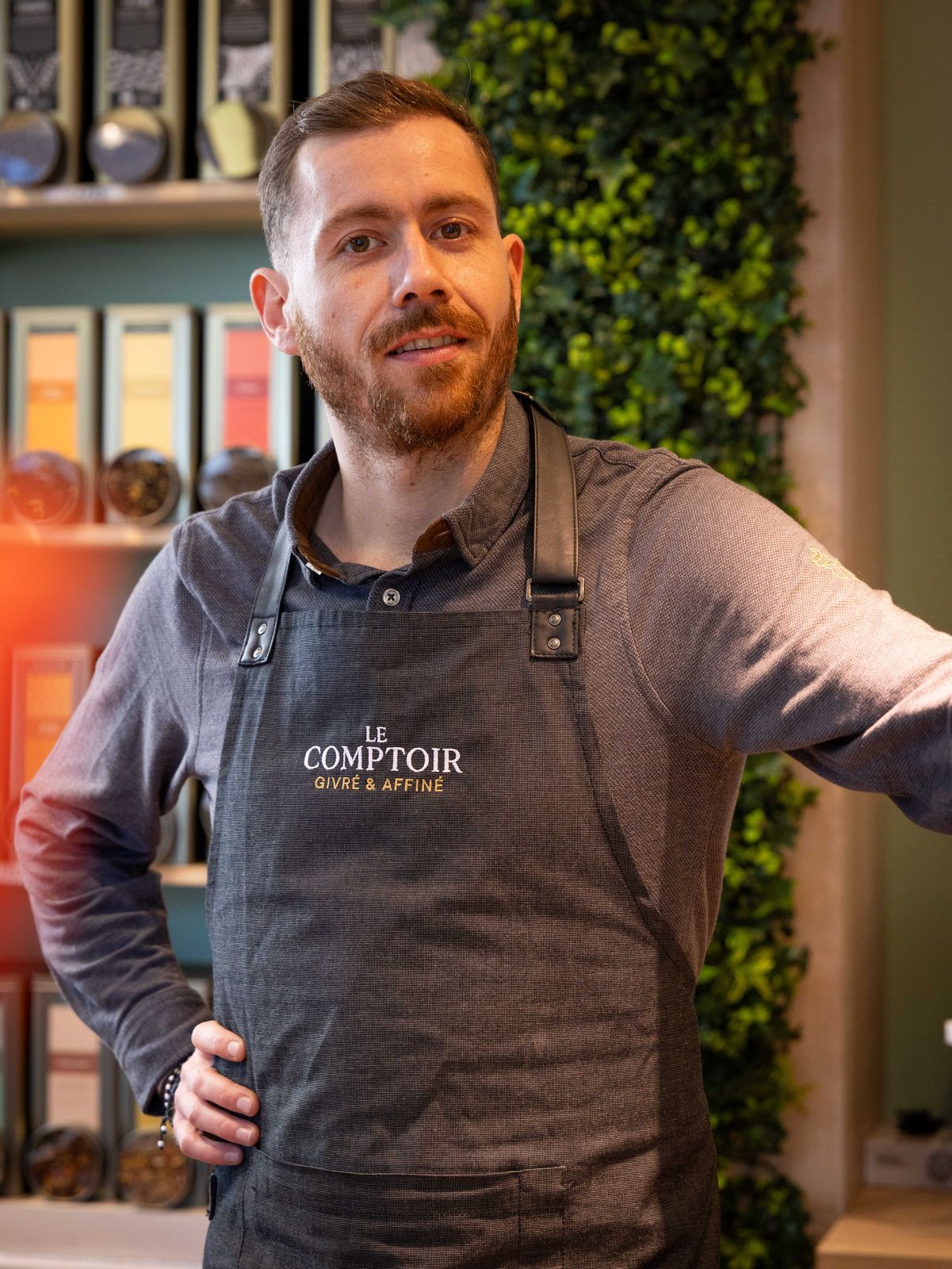 Portrait au Comptoir, en tablier brodé, la main sur la hanche devant le mur de thés